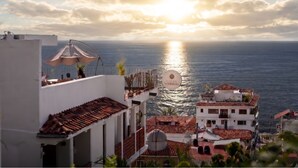 Un edificio costero con balcón que da al mar, un letrero que dice 'RAICES NOMADAS', y un cielo despejado con el sol poniéndose.