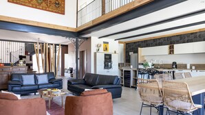 A spacious living area with a black leather sofa, a brown ottoman, and a dining table with wicker chairs. A kitchen with a stone backsplash and modern appliances is visible in the background. - Le Domaine de la Haye - Group gîte for 16 people - 15 minutes from Puy du Fou (Saint-Christophe-du-Bois)