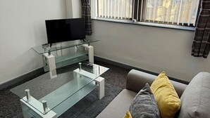 A modern living room with a glass coffee table, a grey sofa with yellow and grey cushions, a flat-screen TV on a glass stand, and a window with blinds overlooking a cityscape.