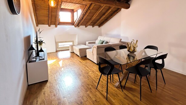 Ein gemütlicher Loftsitzbereich mit Holzbalkendecke, einem Essbereich mit Glastisch und schwarzen Stühlen, einem weißen Sofa mit Kissen sowie einer kleinen weißen Kommode mit Spiegel und Dekoration.