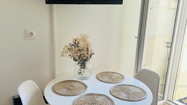 A dining area with a round table, four white chairs, a vase with dried flowers, and a flat-screen TV mounted on the wall. - Apartment triple gare Angers (Angers)