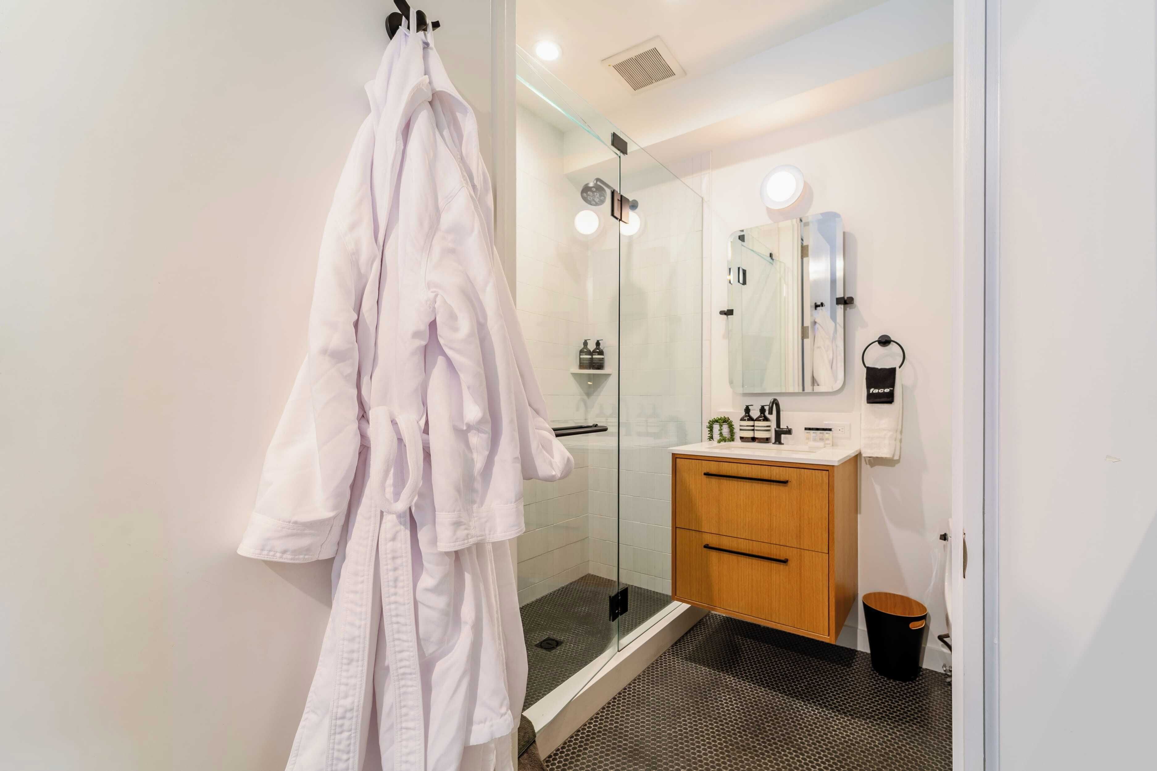 Un baño moderno con cabina de ducha de vidrio, tocador de madera y una bata blanca colgada de un perchero.