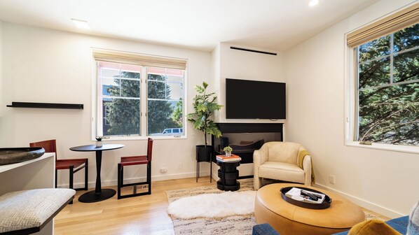 Una sala de estar con sofá, una mesa de centro, televisor, chimenea y ventana con vista a árboles.