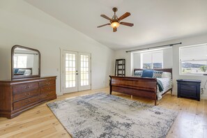 A bedroom with a wooden dresser, a bed with a patterned bedspread, a ceiling fan, and a view through large windows. - Creekside Florrisant Hideaway - Great for Anglers! (Florissant)