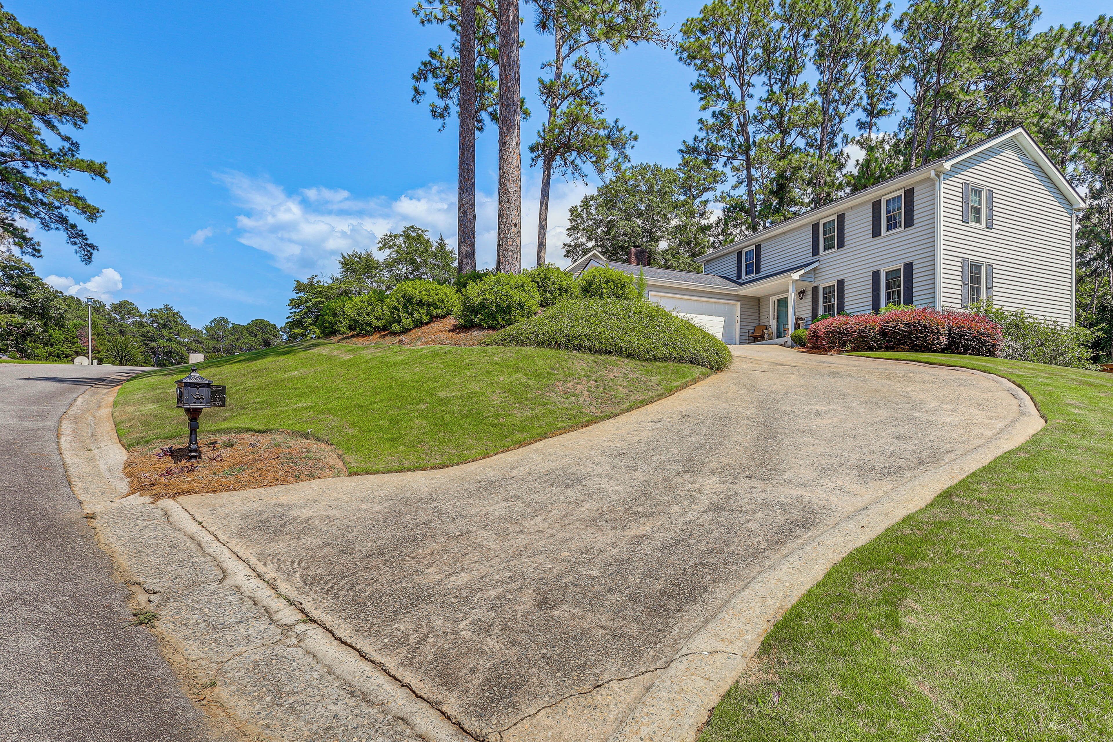 A residential driveway leading to a two-story house with a curved driveway, surrounded by well-maintained lawns and tall trees.