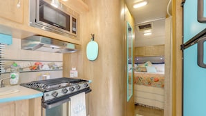A compact kitchen with a stove, microwave, and oven, leading to a bedroom with a bed and curtains. - 6 Mi to Wimberley Square: 'Happy Days Cruiser' (Wimberley)