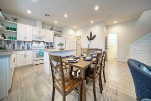A modern kitchen with white cabinetry, a central island with seating, and a dining area with wooden chairs. - Sleeps 20 2 Homes in Mercer Village w/ Pool Heart of Old Town, Walkable (Port Aransas)
