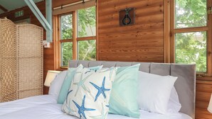A bed with white bedding and starfish-patterned pillows, wooden walls, and large windows with a view of trees. - The Sharpham Boat House (Totnes)