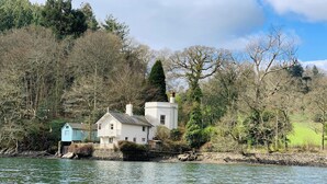 A white house with a balcony by a calm river, surrounded by trees and greenery. - The Sharpham Boat House (Totnes)
