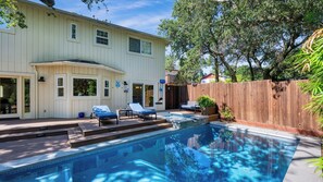 Une piscine avec des chaises longues, une terrasse en bois et une grande maison dotée de nombreuses fenêtres.