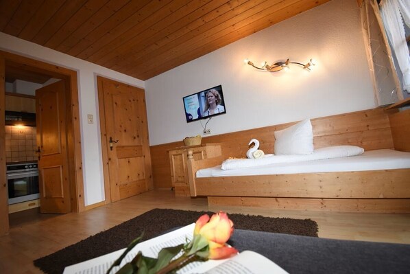 Una habitación acogedora con techo de madera, una cama con ropa de cama blanca, un televisor montado en la pared y una cocina visible a través de una puerta abierta.