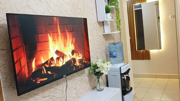 A modern living room with a flat-screen TV displaying a fireplace, a water dispenser, and a small cabinet with a plant. - one bedroom apartment in juja (Juja)