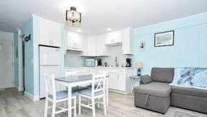 Un espacio habitable compacto con kitchenette, una zona de comedor con mesa y sillas, un sofá gris y una pequeña alfombra.