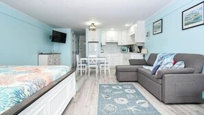 Una habitación con una cama, un sofá gris, una mesa de comedor con sillas, una zona de cocina y un televisor montado en la pared.