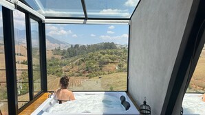 Une personne se détend dans un bain à remous, profitant d’une vue panoramique sur les collines et les arbres, grâce à de grandes fenêtres.
