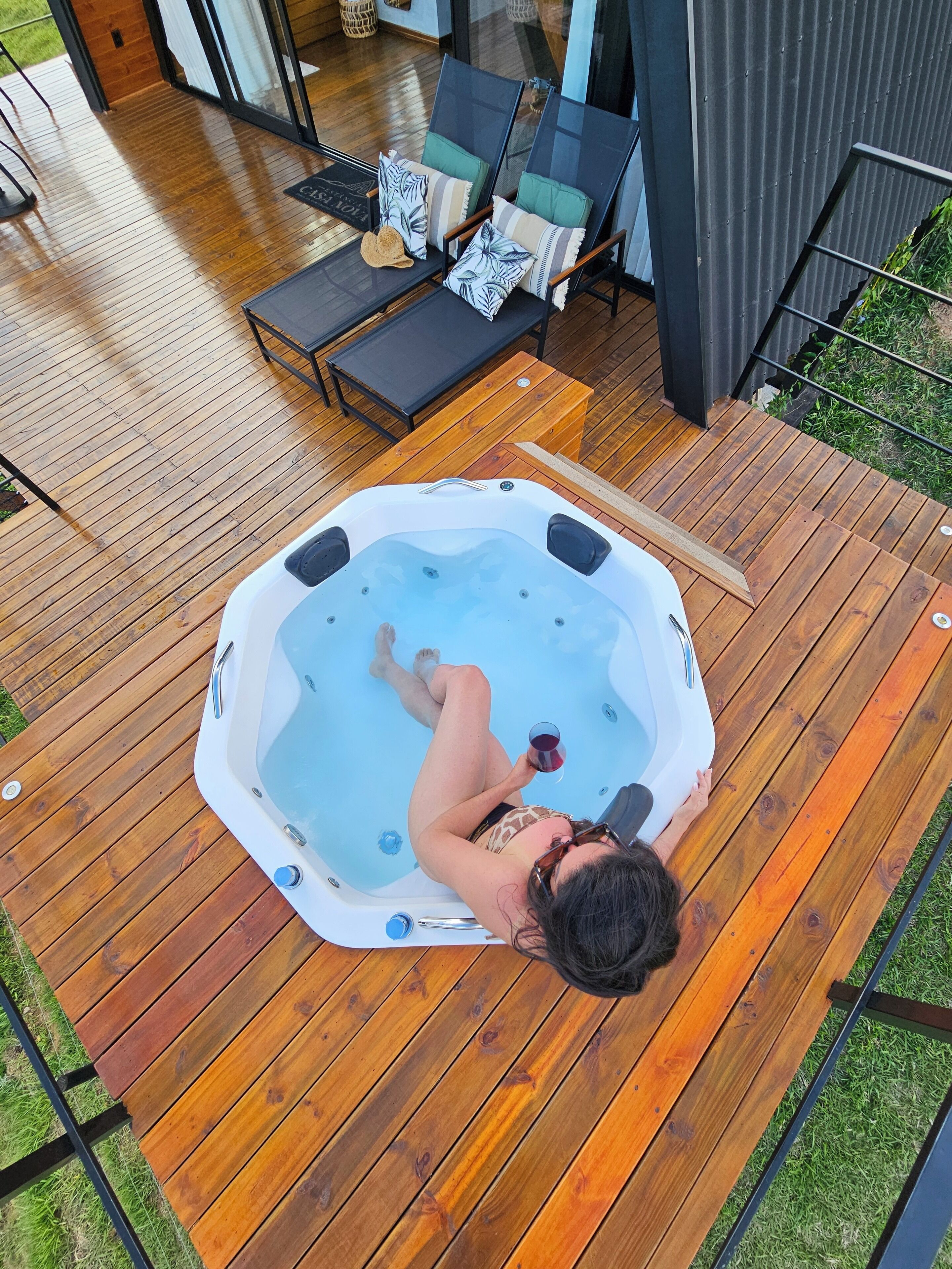 Une personne se détend dans un bain à remous sur une terrasse en bois, avec des chaises longues et une porte vitrée en arrière-plan.