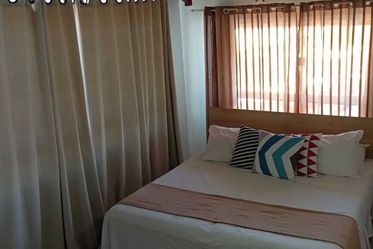 A bed with a wooden headboard, beige curtains, and a bedside table with a lamp.