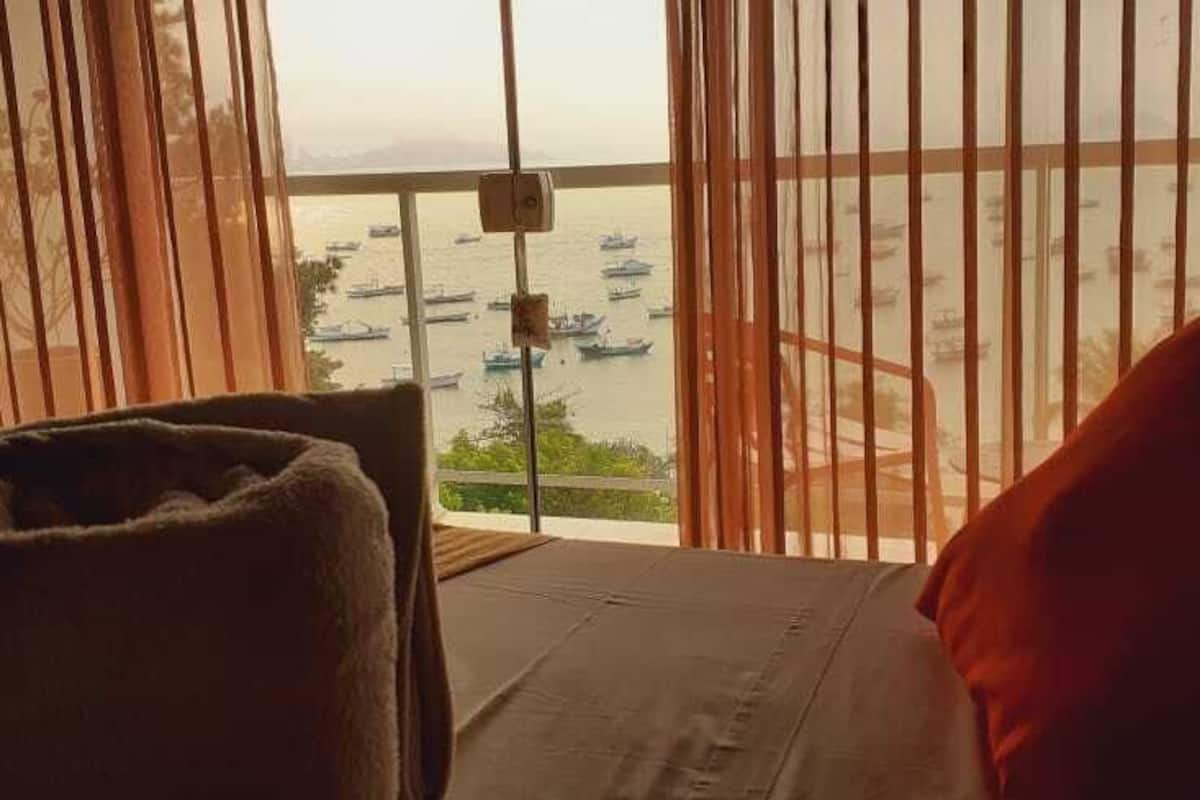 A room with a bed, a chair, and a view of boats on the water through large windows.