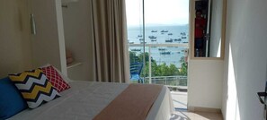 A hotel room with a bed, a view of the sea with boats, and a balcony with a railing. - Maricá Apart Hotel (Penha)