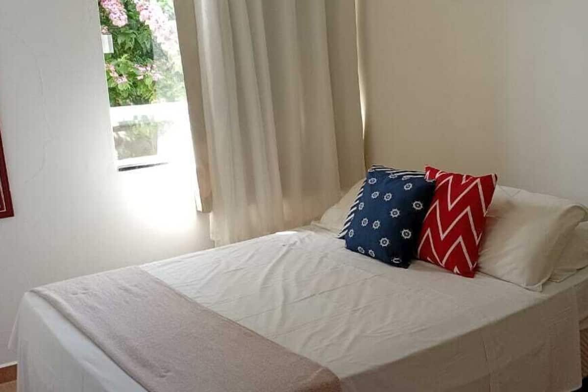 A neatly made bed with a white bedspread and two pillows, one with a blue pattern and one with a red zigzag pattern, in a room with a window and a shelf with folded towels.