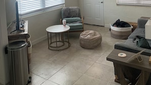 Una sala de estar con ventilador de techo, una puerta con ventanal decorativo, una mesa redonda, un banco acolchado y un cubo de basura.