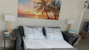 Un dormitorio con una cama, dos mesitas de noche, un cuadro de un atardecer y una lámpara en cada mesita.