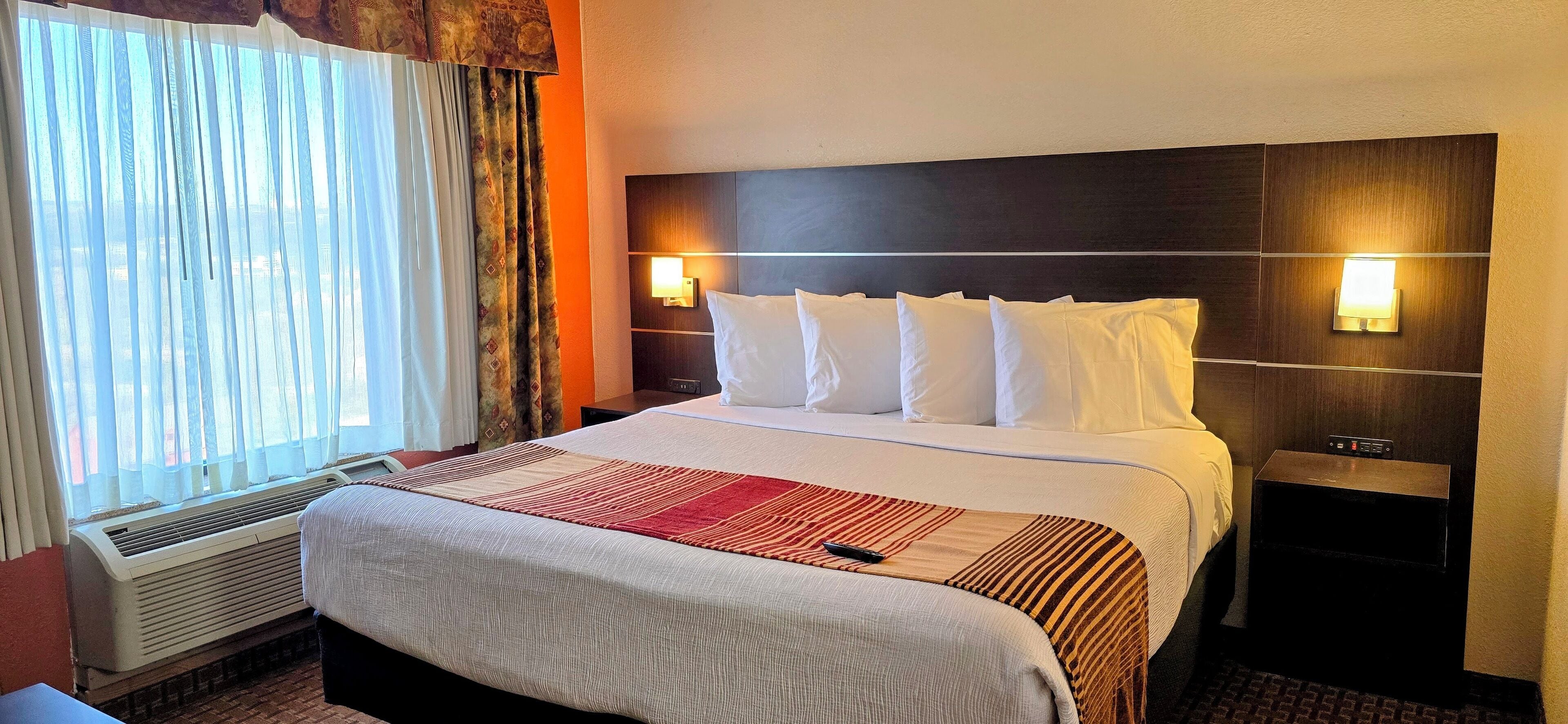A hotel room with a large bed, a bedside table, a wall-mounted lamp, and a window with curtains.