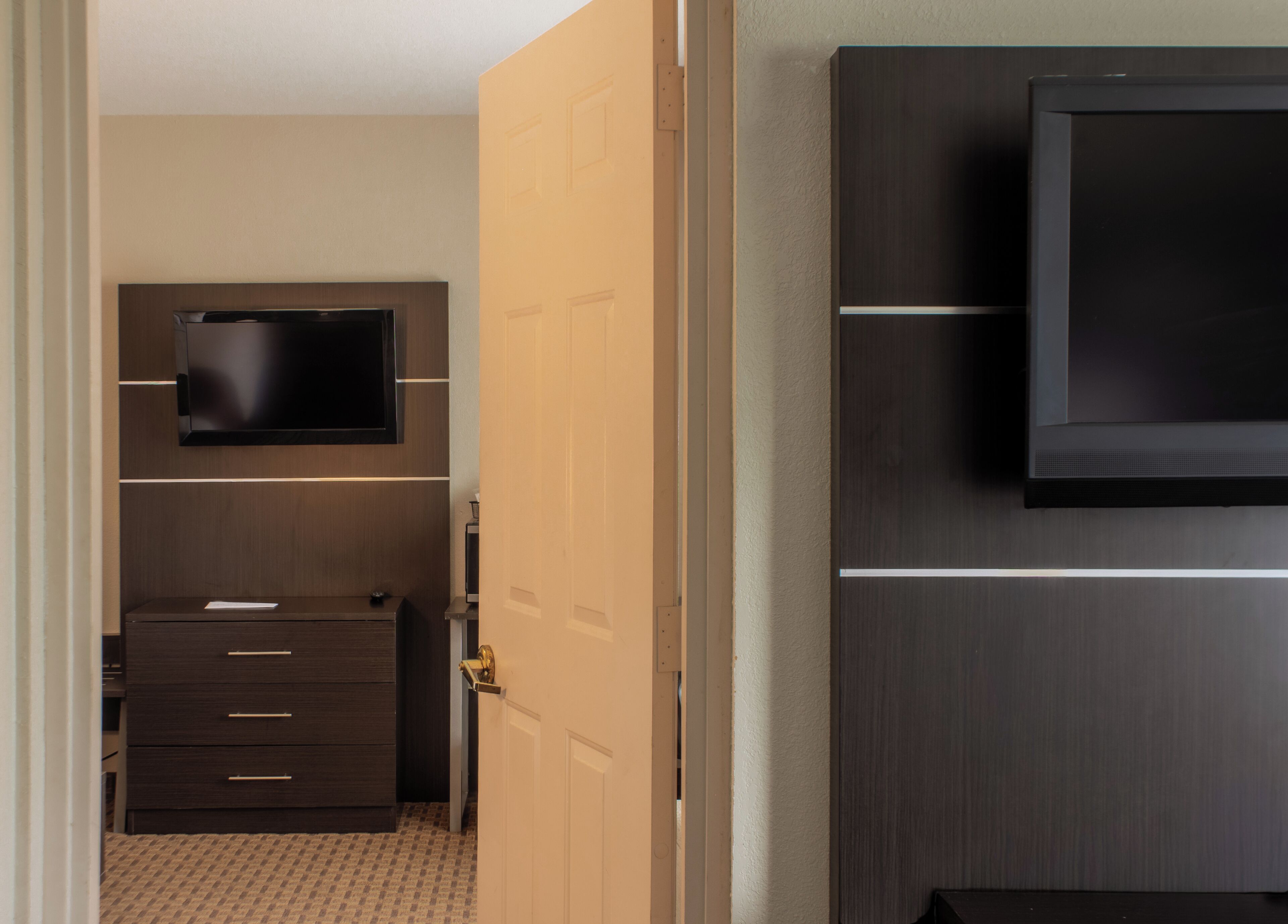 A hotel room with a flat-screen TV mounted on the wall, a wooden dresser with drawers, and a door leading to another room.