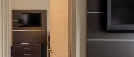 A hotel room with a flat-screen TV mounted on the wall, a wooden dresser with drawers, and a door leading to another room.