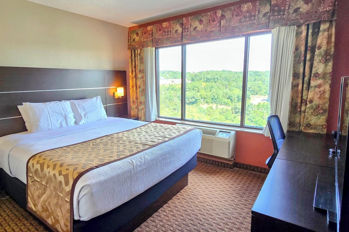 Ein Hotelzimmer mit einem großen Bett, Nachttischlampen, einem Schreibtisch mit Stuhl und einem Fenster mit Blick auf Grünflächen.