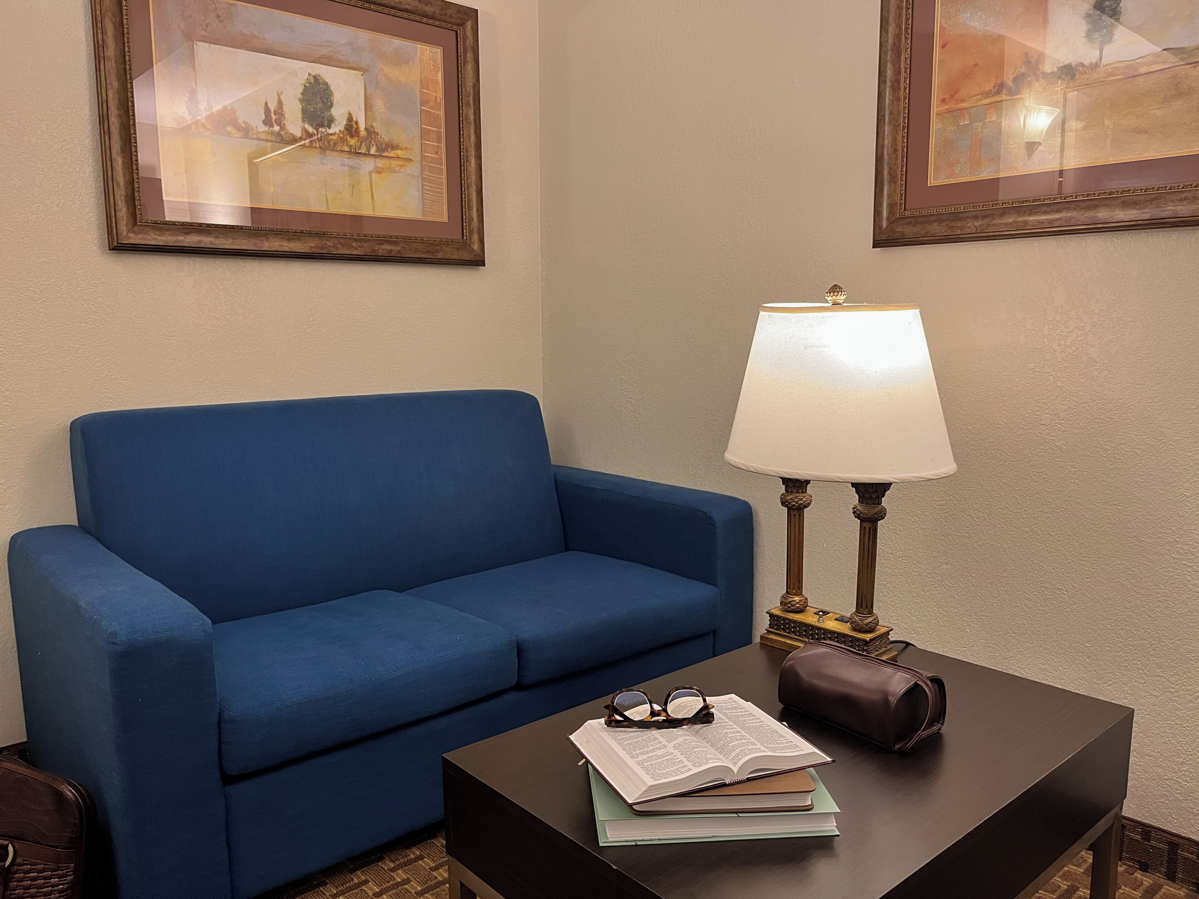 A room with a blue sofa, a table with a lamp, and two framed pictures on the wall.