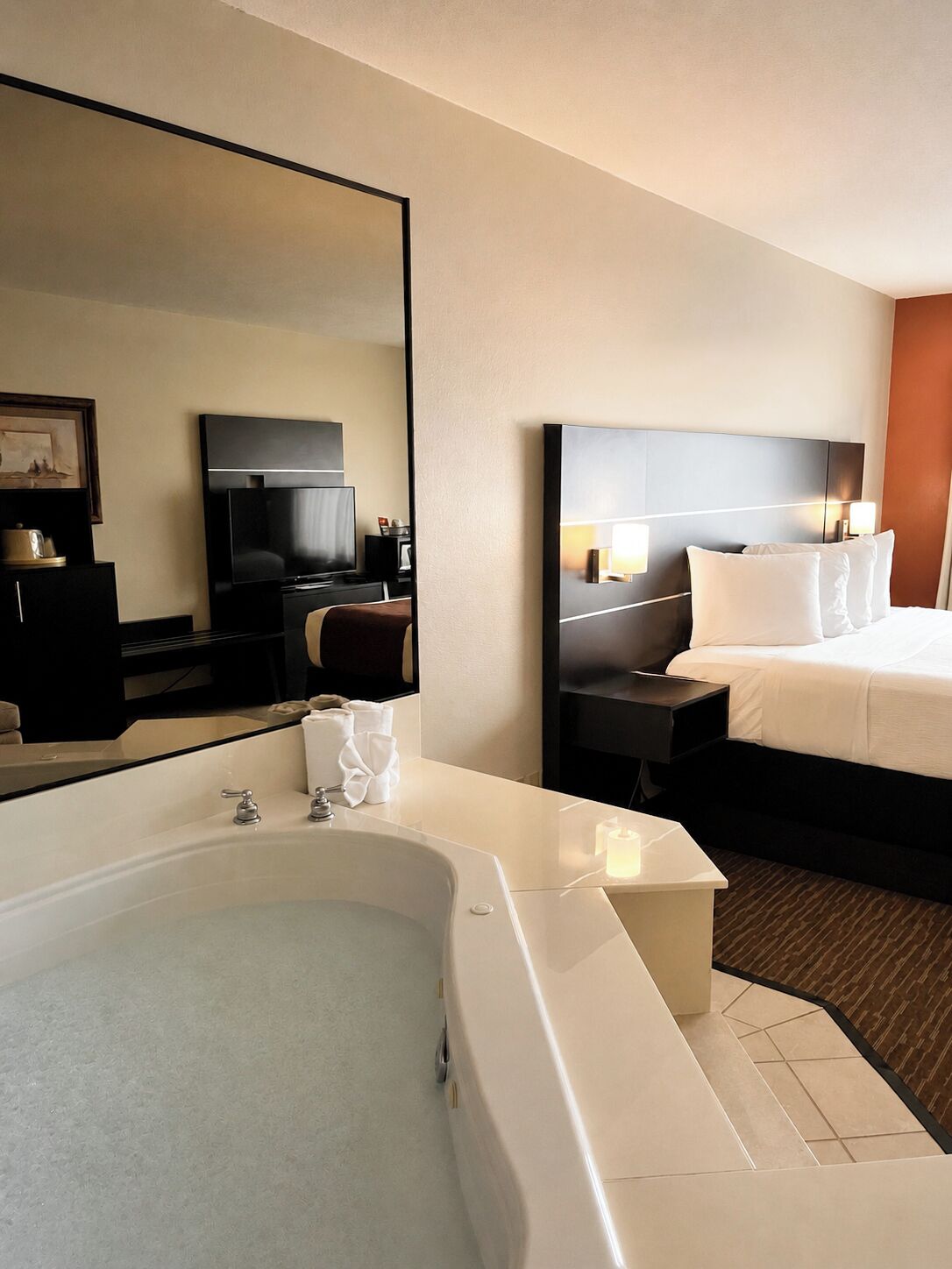 Habitación de hotel con una bañera grande, cama con ropa blanca, televisor de pantalla plana y un espejo.