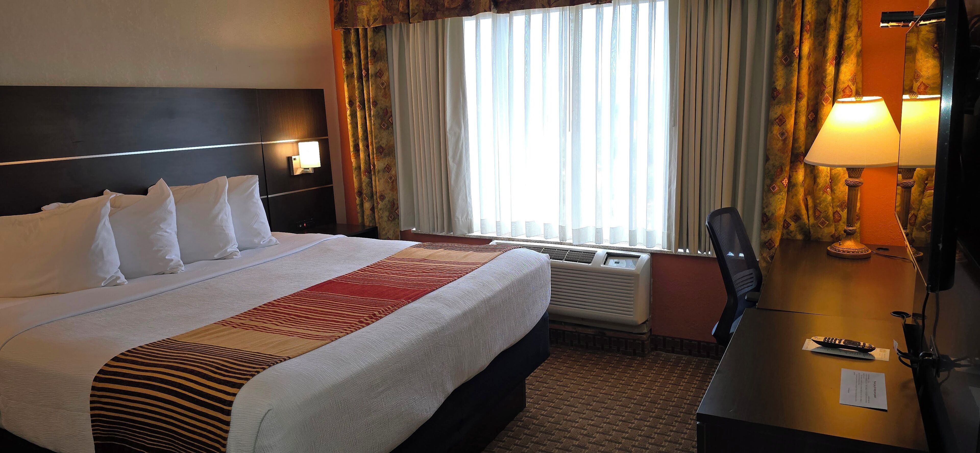 Una habitación de hotel con una cama grande, un escritorio con lámpara y una ventana con cortinas.