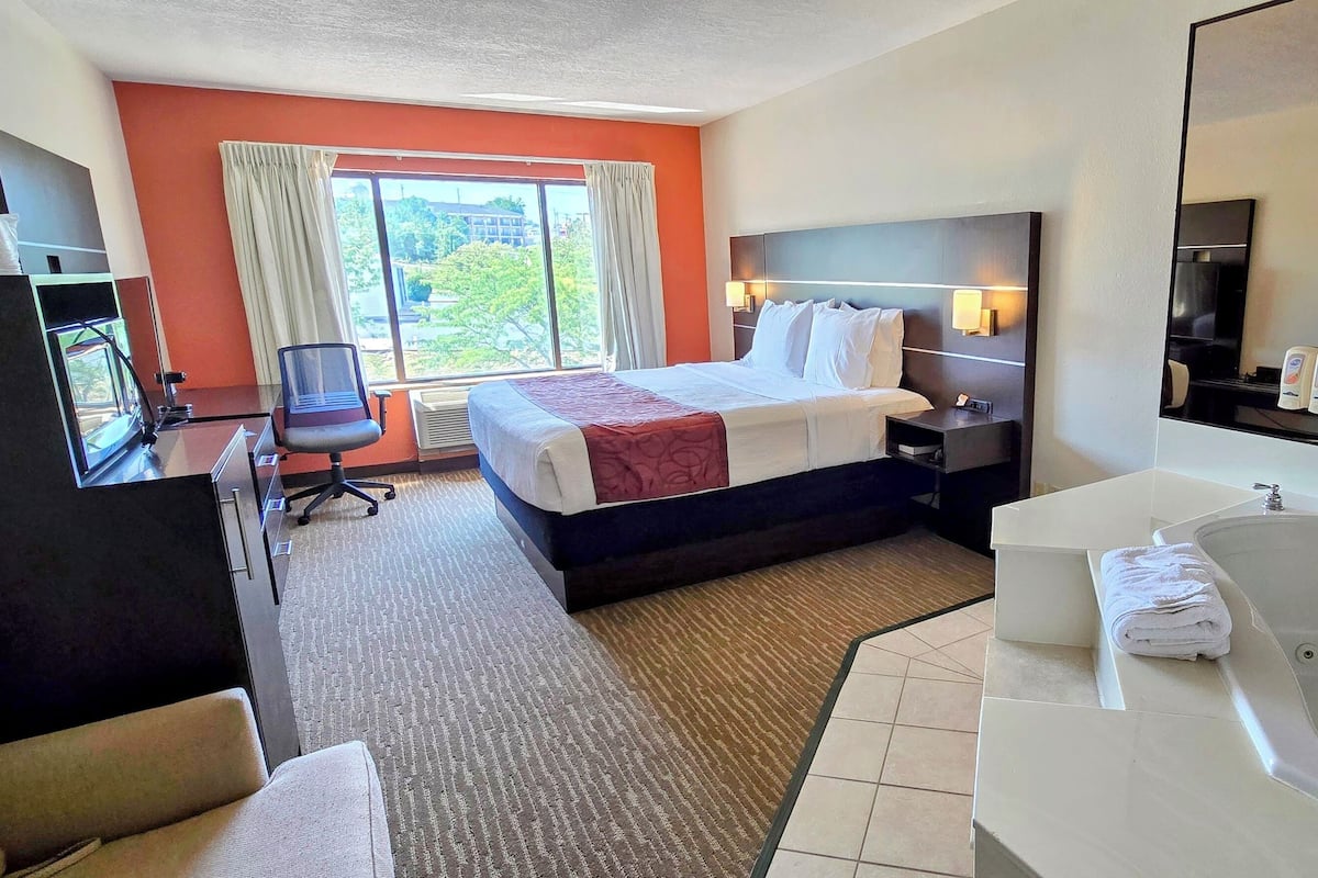 Ein Hotelzimmer mit einem großen Bett, einem Schreibtisch mit Computer, einem separaten Sessel, einem großen Fenster mit Vorhängen, einem Badezimmer mit Badewanne und Waschbecken sowie einem Spiegel.