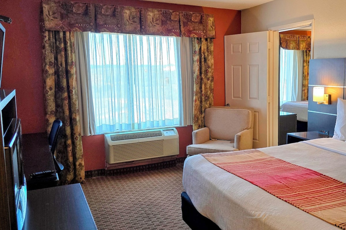 Ein Hotelzimmer mit einem großen Bett, einem Schreibtisch, einem Stuhl und einem fensterbelaubten Fenster.