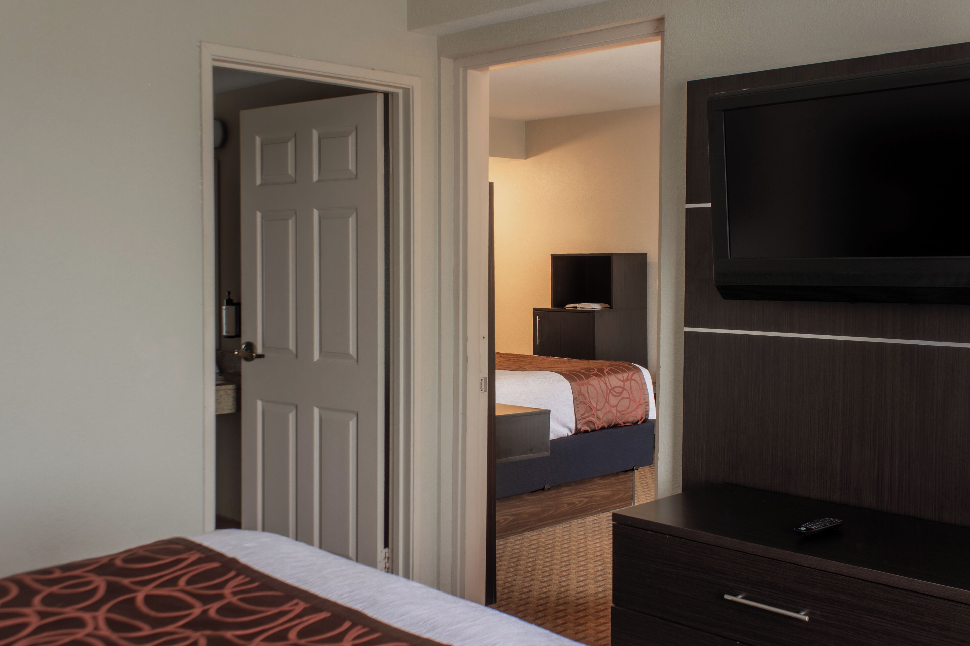 A hotel room with a bed, a TV mounted on the wall, a dresser, and a door leading to another room.