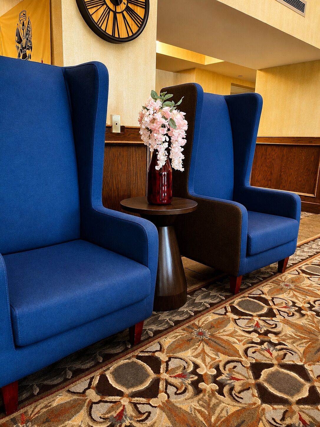 Un hall d’hôtel avec deux fauteuils bleus, une petite table ronde ornée d’un vase de fleurs et un tapis à motifs.