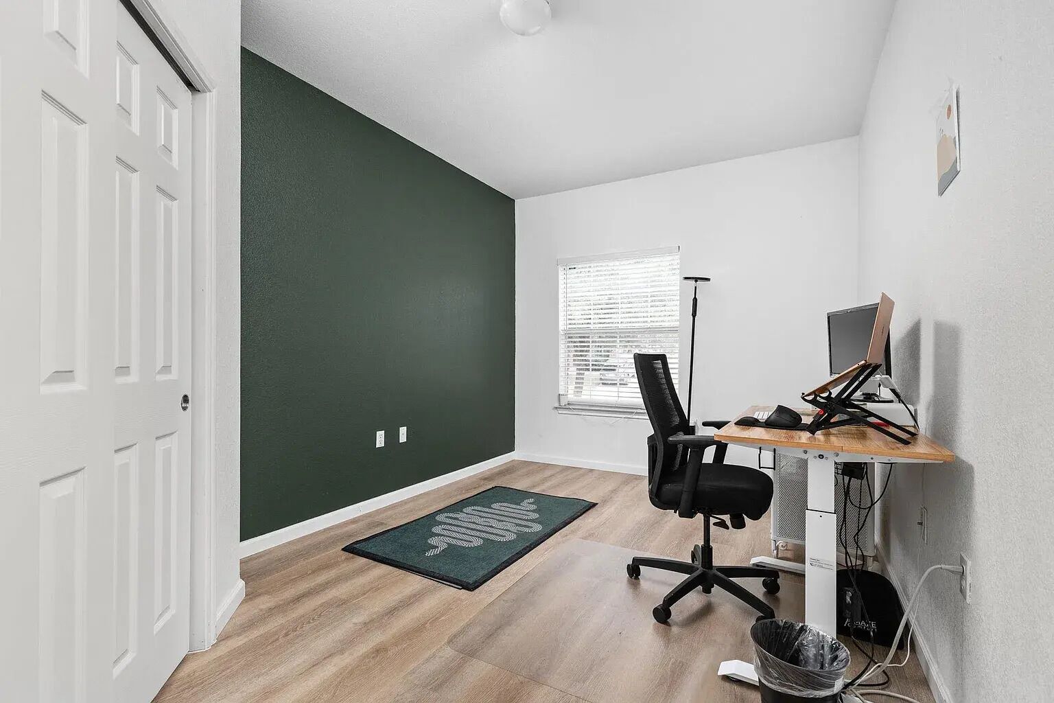 A room with a green accent wall, a wooden desk with a computer, a black office chair, and a window with blinds.
