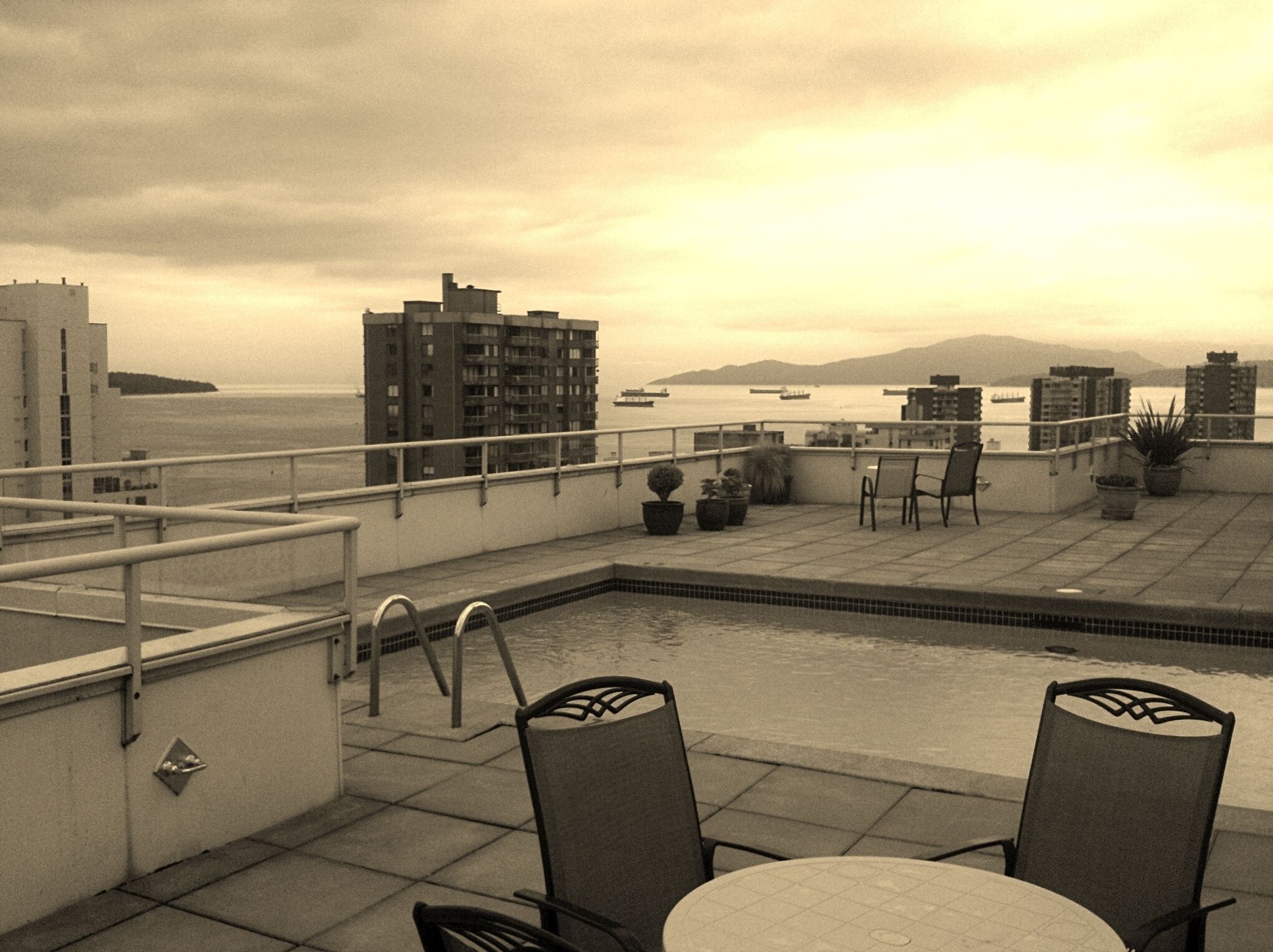 Une terrasse sur le toit avec une piscine, des chaises, des plantes en pot et une vue sur l’océan et les bâtiments éloignés.