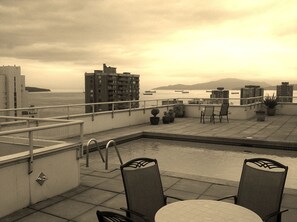 Terraza en la azotea con piscina, sillas, plantas en macetas y vistas al océano y edificios al fondo.