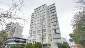 A multi-story residential building with balconies, a parking lot, and a street view.