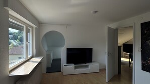 A modern living room with a large window, a flat-screen TV on a white entertainment unit, and a grey rug on the wooden floor.