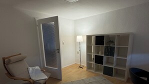 A living room with a wooden floor, a grey rug, a fluffy rug, a chair, a small table, a lamp, and a shelving unit.