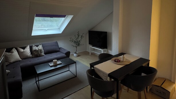 A modern loft apartment with a high ceiling, a skylight, a sofa, a coffee table, a TV, a dining table with chairs, and a small plant.