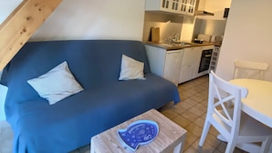 A cozy living area with a blue sofa, a small wooden table, and a white dining table with chairs. A kitchen with white cabinets and a stove is visible in the background. - Small house Hourtin port (Hourtin)