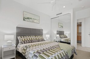 Ein Schlafzimmer mit grauem, gepolsterten Kopfteil, einer gesteppten Bettdecke, Nachttischlampen und einem großen Spiegel.