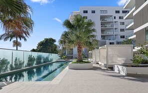 Ein moderner Poolbereich im Freien mit klarem Wasser, umgeben von Palmen und einem weißen Gebäude mit Balkonen.