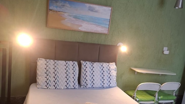 A neatly made bed with a patterned pillow set, a painting of a beach scene, and a small table with chairs in a room with green walls. - Aconchego Volta Redonda 449 (Volta Redonda)