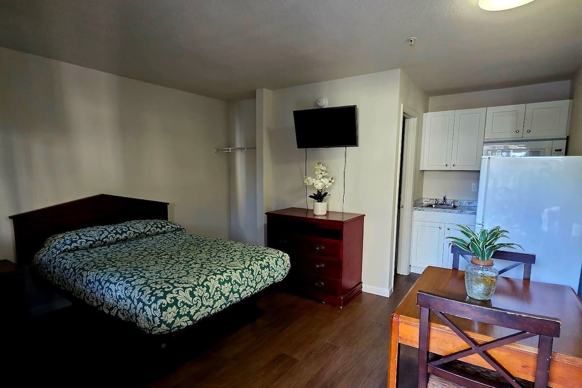 A compact room with a bed, a desk with a chair, a television, and a kitchenette.