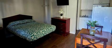 A compact room with a bed, a desk with a chair, a television, and a kitchenette.